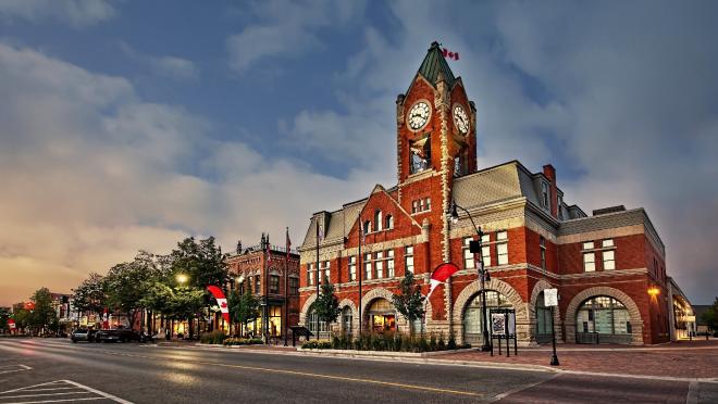 A historic red-brick building with a clock tower, Canadian flags, and arched windows, illuminated against a twilight sky on a quiet downtown street
