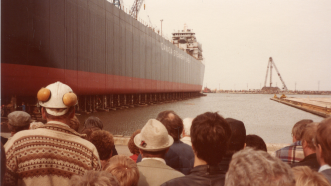 a group of people stand facing a large ship launching into the harbour