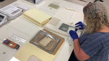 A woman with curly hair sits at a table with gloves on looking through collections materials.