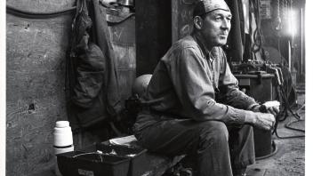 A shipyard worker sits on a bench in a workshop. An open lunch pail and thermos is on the bench beside him.