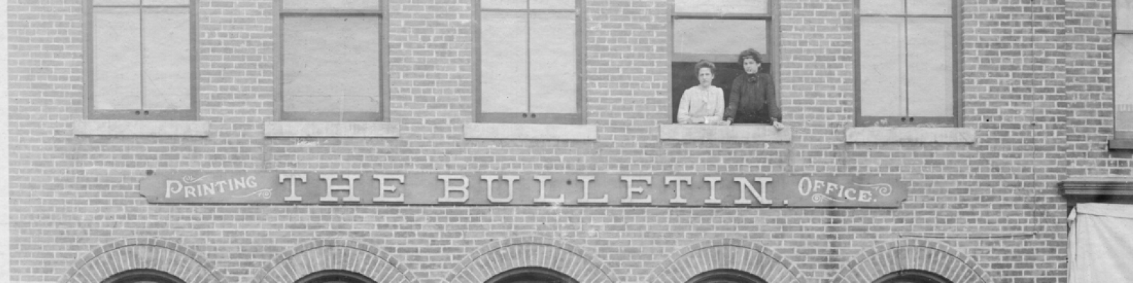 Black and white image of the old Enterprise Bulletin building with two women in 1800 clothes at the window.