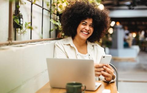 Woman smiling. They are typing on their cellphone and have a laptop computer in front of them.